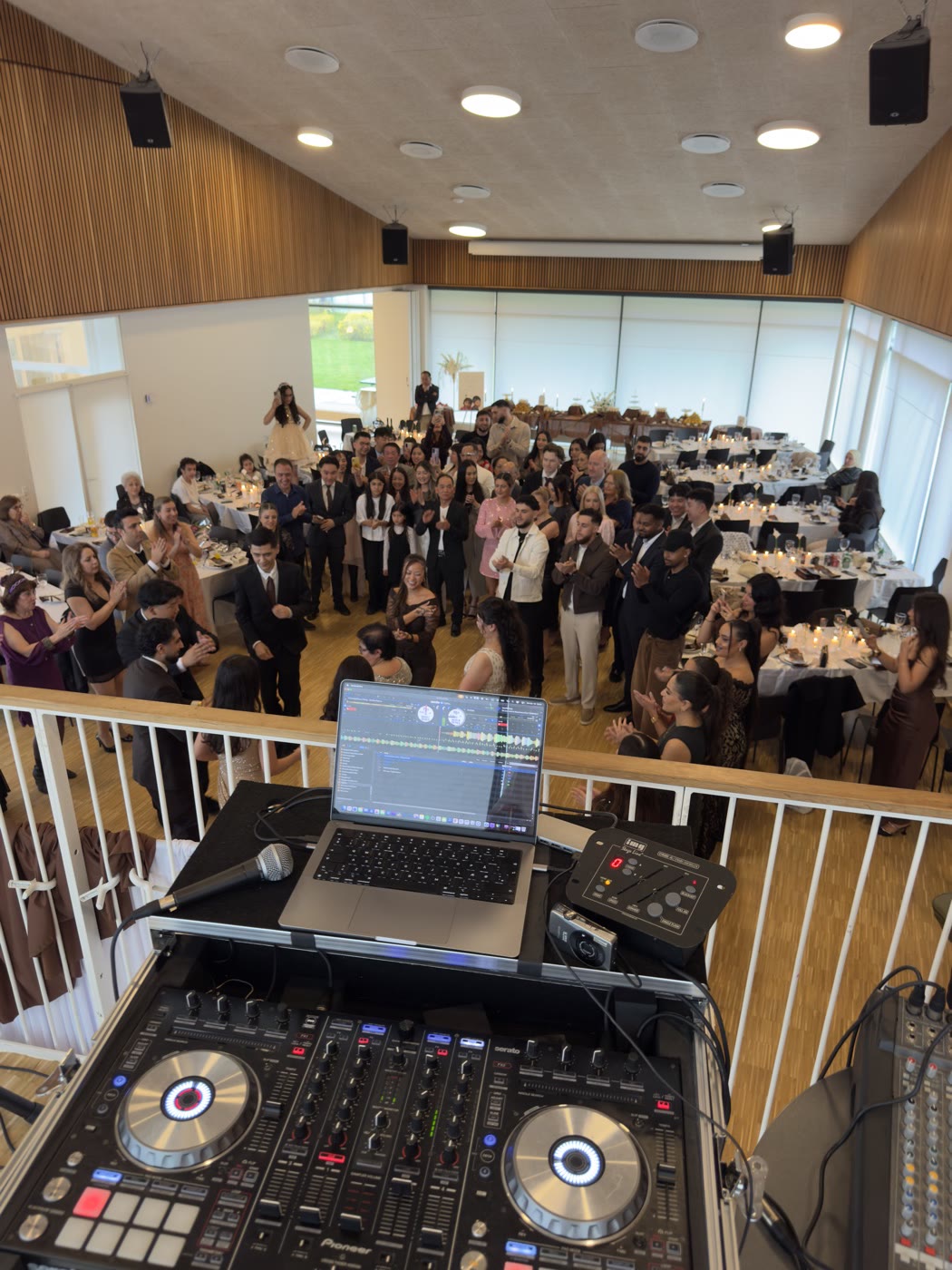 DJ-pult med mikrofon på en balkon over en festsal fuld af gæster ved dækkede borde